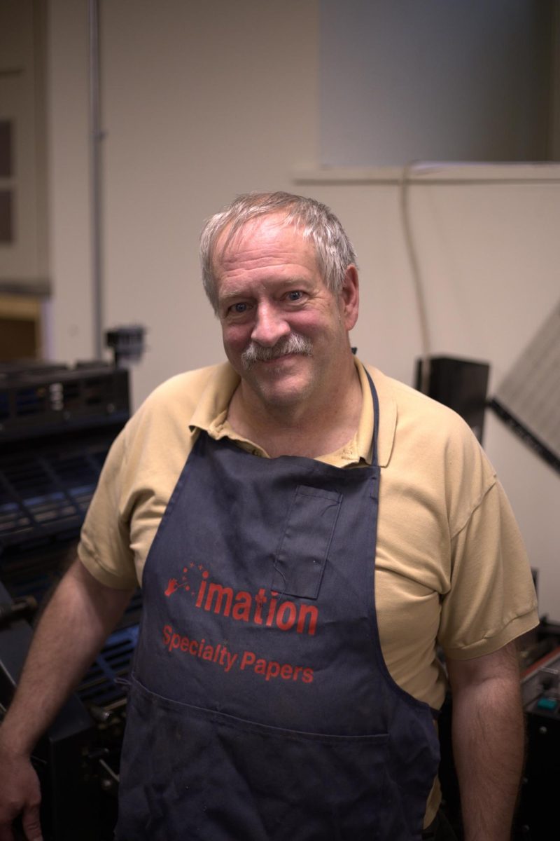  Keith Morse, manager of the print shop, smiles next to the printing press wearing his favorite apron.